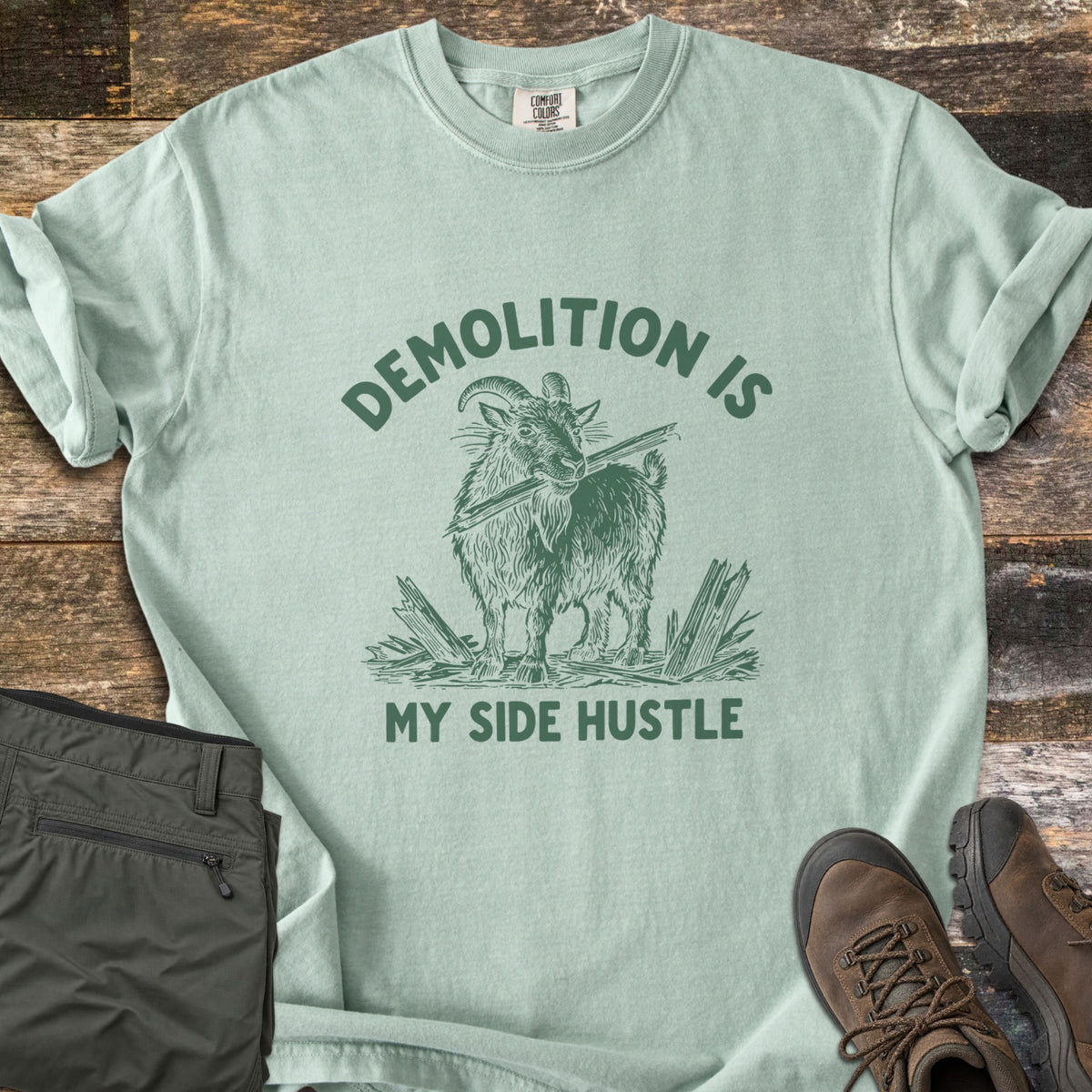 Demolition Is My Side Hustle Comfort Colors T-shirt in Bay, laid flat. Green print of a shaggy goat holding a broken board in its mouth among splintered wood, with the words Demolition Is arched above and My Side Hustle below. Olive pants and brown hiking boots lie on weathered wood.