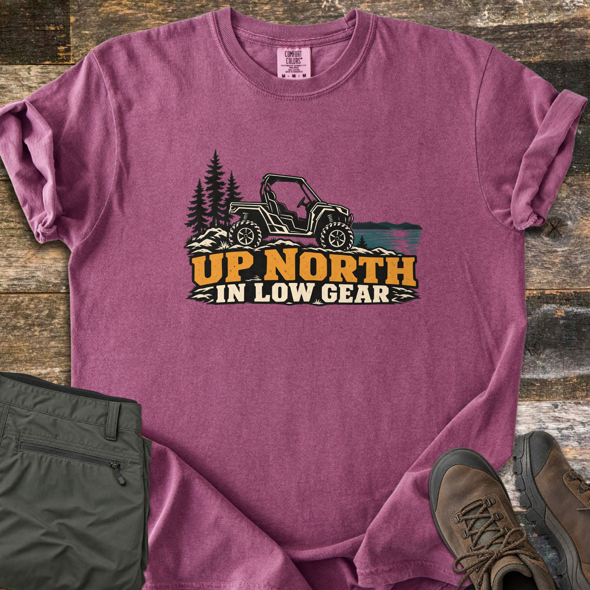 Up North in Low Gear Comfort Colors T-shirt in Berry, laid flat. A side-by-side off-road vehicle sits on rocks by pine trees and a lake above the words Up North in Low Gear. Olive cargo pants and brown hiking boots lie beside it on rustic wood planks.