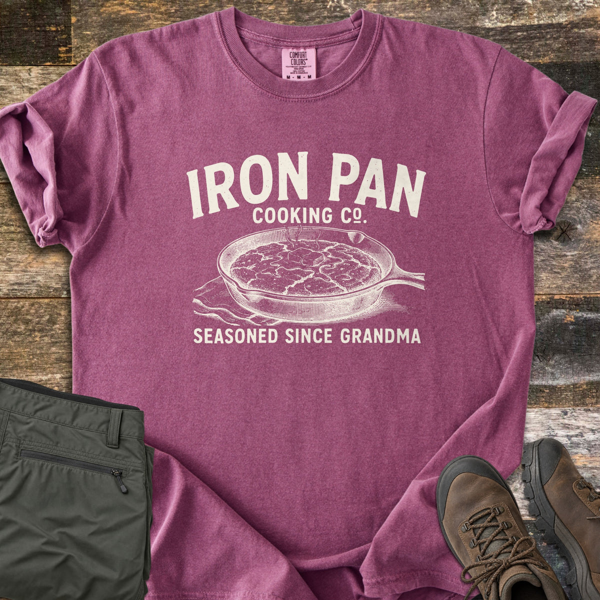 Iron Pan Cooking Co Comfort Colors T-shirt in Berry, laid flat. Cream print of a cast iron skillet of cornbread with melting butter on a napkin, under the words Iron Pan Cooking Co. and above Seasoned Since Grandma. Folded olive pants and brown hiking boots lie on rustic wood.