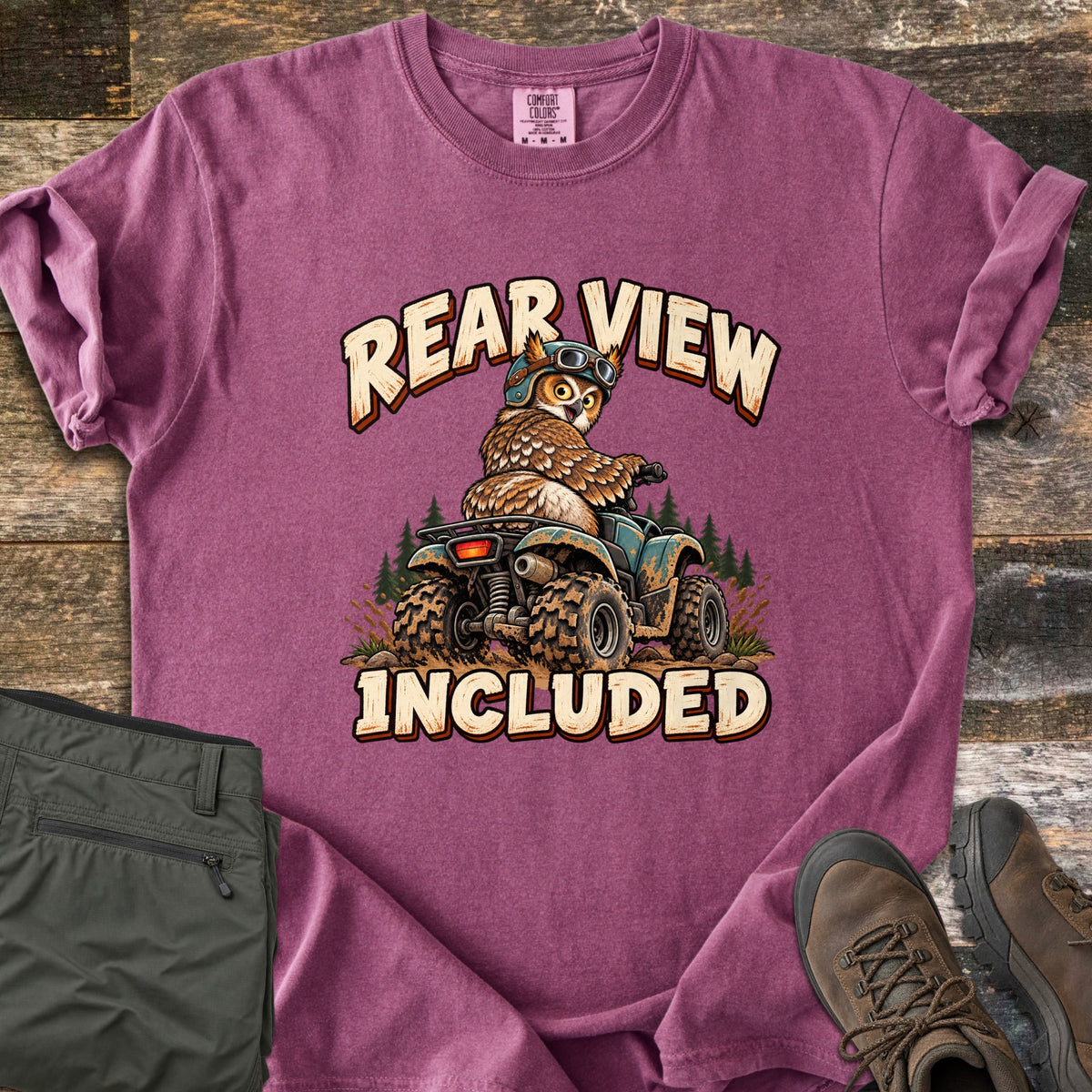 Rear View Included Comfort Colors T-shirt in Berry, laid flat. A cartoon owl in a helmet and goggles rides a muddy ATV past pine trees, looking back over its shoulder, between the words Rear View Included. Olive cargo pants and brown hiking boots lie on a wood floor.