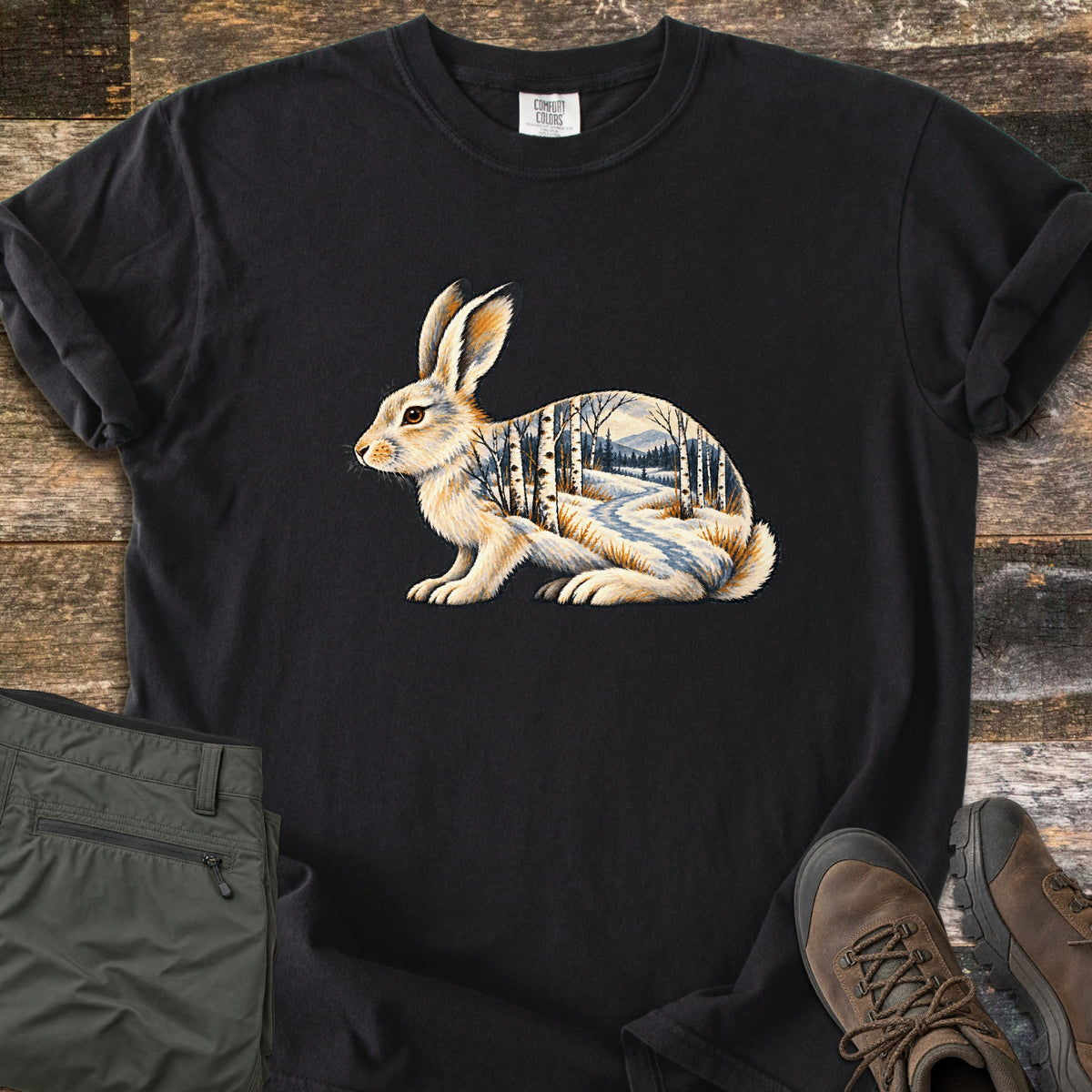 Snowshoe Hare Birch Woods Comfort Colors T-shirt in Black, laid flat. A cream snowshoe hare sits in profile, its body filled with a snowy birch forest, a winding path and distant mountains; no words. Sleeves rolled, on weathered wood with olive pants and brown hiking boots.