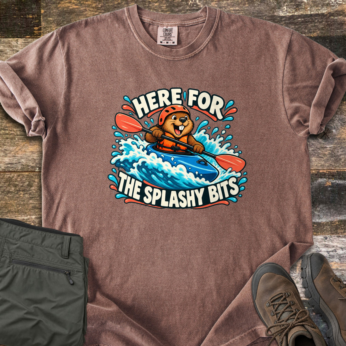 Splashy Bits Comfort Colors T-shirt in Espresso, laid flat. A cartoon beaver in an orange helmet and life vest paddles a blue kayak through whitewater, framed by the words Here For The Splashy Bits. Olive pants and brown hiking boots lie beside it on weathered wood.