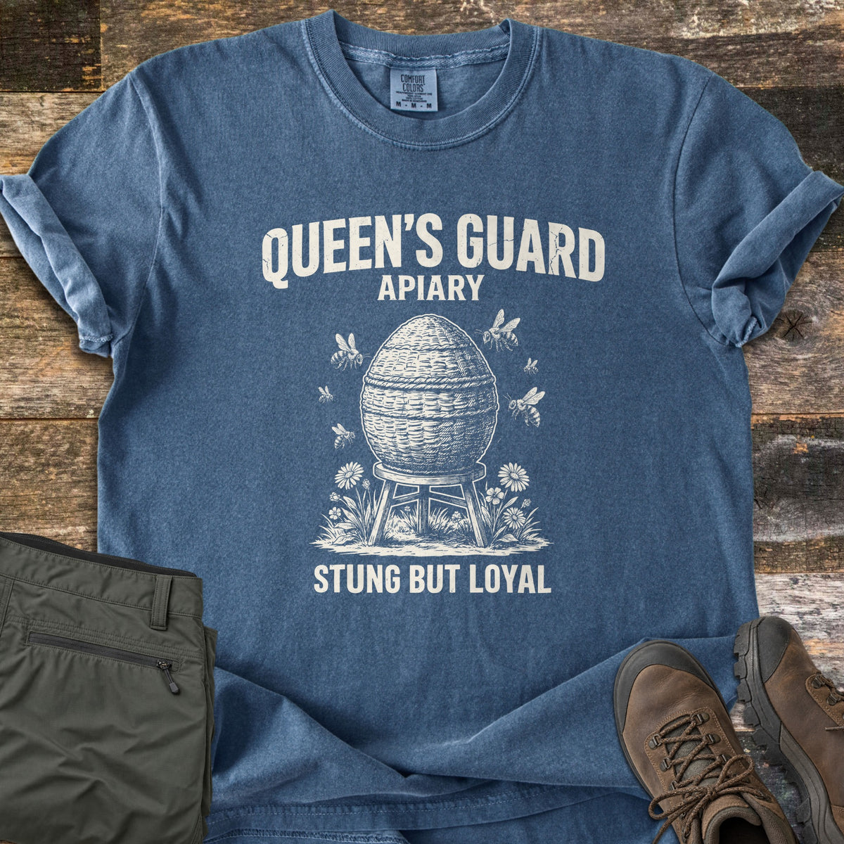 Queens Guard Apiary Comfort Colors T-shirt in Denim, laid flat. A woven skep beehive on a wooden stand among wildflowers, with bees flying around it, under the words Queen's Guard Apiary and above Stung But Loyal. Olive cargo pants and brown hiking boots lie on weathered wood.