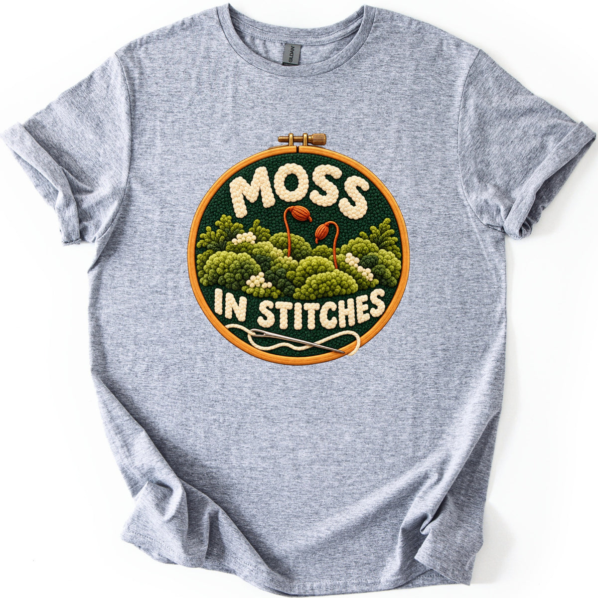 Moss in Stitches Comfort Colors T-shirt in Sport Grey, laid flat. A wooden embroidery hoop frames stitched green moss with two orange spore capsules on stalks, cream yarn letters reading MOSS IN STITCHES, and a threaded needle. Sleeves rolled, on a white background.