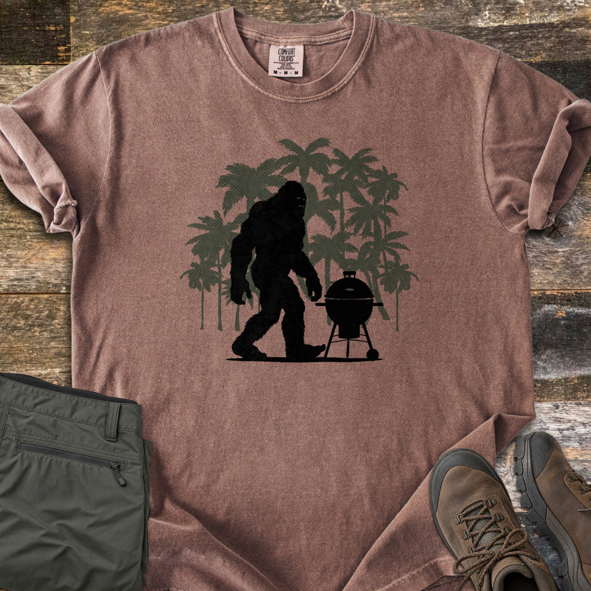 Squatch BBQ Smoker Comfort Colors T-shirt in Espresso, laid flat. A black Bigfoot silhouette walks past a kettle grill on wheels, set against dark green palm tree silhouettes; no words are printed. The shirt lies on weathered wood beside olive pants and brown hiking boots.