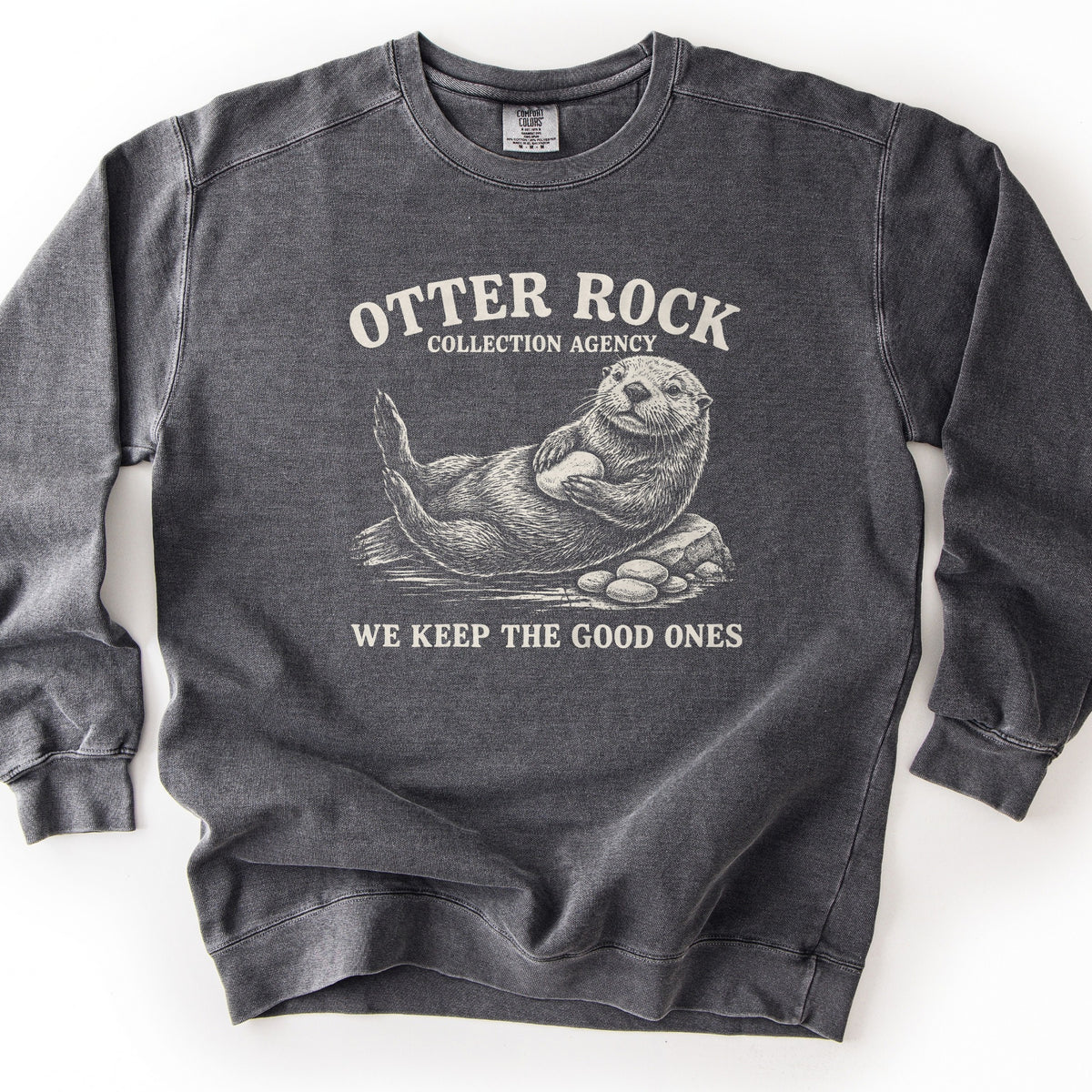 Otter Rock Collection Agency Comfort Colors crewneck sweatshirt in Pepper, laid flat. A cream sketch-style sea otter floats on its back clutching a round stone, beside a rock and a pile of pebbles, between the words Otter Rock Collection Agency and We Keep the Good Ones. Shown on a white background.