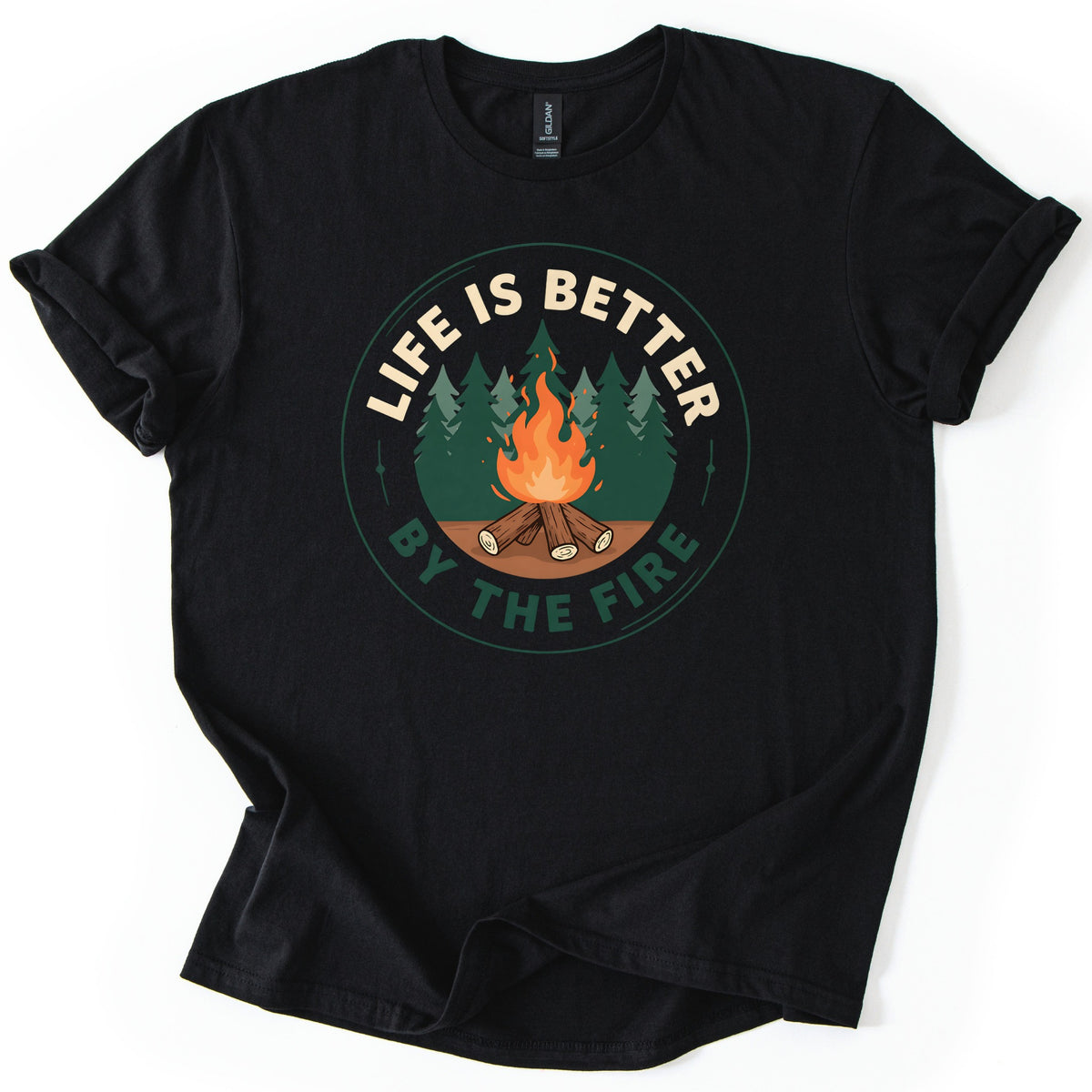 Life Is Better By The Fire Badge Comfort Colors T-shirt in Black, laid flat. A round badge shows orange flames rising from stacked logs before a dark green pine forest, with LIFE IS BETTER in cream letters on top and BY THE FIRE in green underneath. The shirt lies on a white surface.