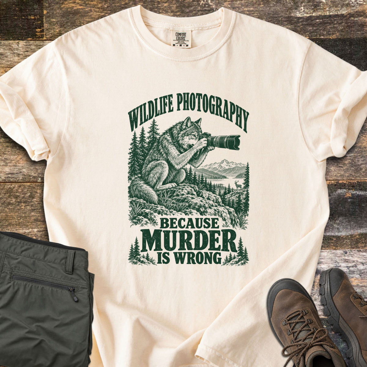 Wildlife Photography Because Murder Is Wrong Comfort Colors T-shirt in Ivory, laid flat. Dark green ink shows a wolf on a rocky ledge aiming a long-lens camera at a distant deer by a mountain lake, under Wildlife Photography and above Because Murder Is Wrong. Olive pants and brown hiking boots lie on dark wood.