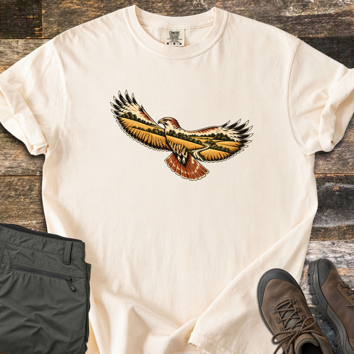 Hawk Over Golden Fields Comfort Colors T-shirt in Ivory, laid flat. A hawk with outstretched wings and a rust-red tail, its wings filled with golden fields, green trees and a winding path; no words. Folded olive hiking pants and brown hiking boots sit beside it on weathered wood.