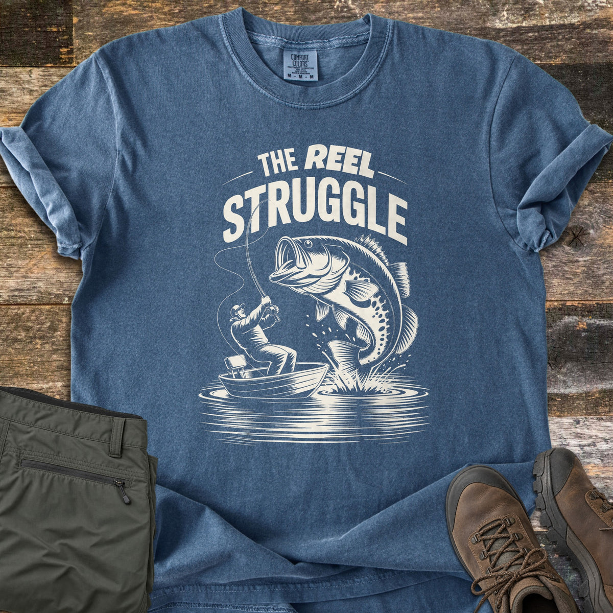 Reel Struggle Comfort Colors T-shirt in Denim, laid flat. A white print of a man in a small boat reeling in a leaping bass, under the words The Reel Struggle. Sleeves are rolled on a wood plank surface beside folded olive pants and brown hiking boots.
