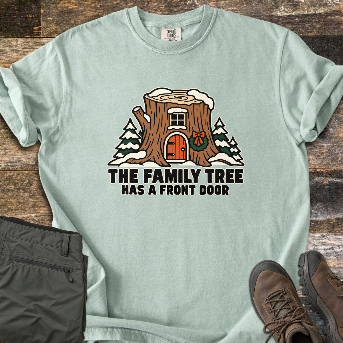 The Family Tree Has a Front Door Comfort Colors T-shirt in Bay, laid flat. A snowy tree stump house with a window, an orange arched door, and a wreath with a bow sits between pine trees above the words The Family Tree Has a Front Door. Olive pants and brown hiking boots lie on dark wood.