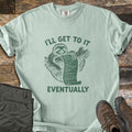 I'll Get To It Eventually Comfort Colors T-shirt in Bay, laid flat. A green sketch of a smiling sloth hugging a branch and holding a long scroll list, between the words I'll Get To It Eventually. Folded olive pants and brown hiking boots sit on a dark wood surface.