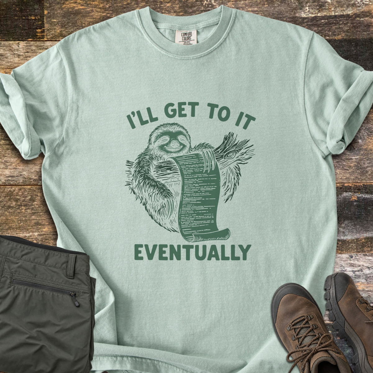 I'll Get To It Eventually Comfort Colors T-shirt in Bay, laid flat. A green sketch of a smiling sloth hugging a branch and holding a long scroll list, between the words I'll Get To It Eventually. Folded olive pants and brown hiking boots sit on a dark wood surface.
