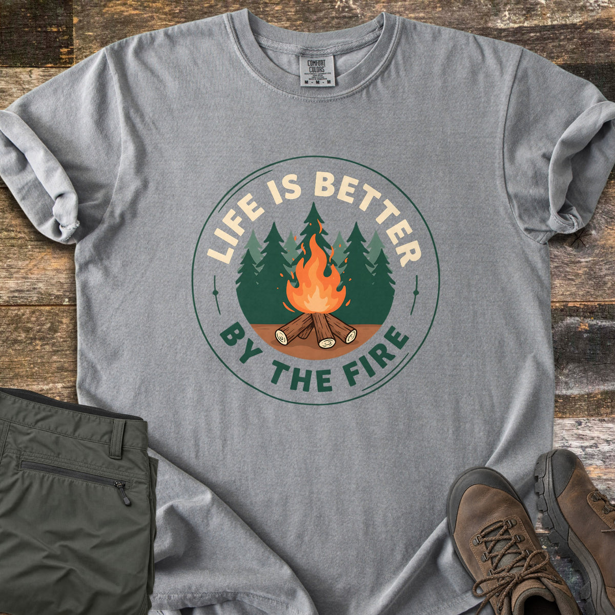 Life Is Better By The Fire Badge Comfort Colors T-shirt in Grey, laid flat. A round badge shows a campfire of crossed logs before pine trees, with LIFE IS BETTER arched above and BY THE FIRE below. Olive hiking pants and brown hiking boots sit beside it on weathered wood planks.