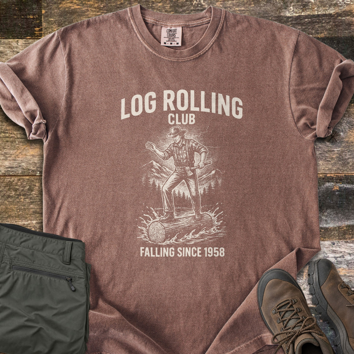 Log Rolling Club Comfort Colors T-shirt in Espresso, laid flat. A sketched lumberjack in a hat and plaid shirt, holding an axe, balances on a log splashing through water below the words Log Rolling Club and above Falling Since 1958. Olive pants and brown boots sit on wood planks.