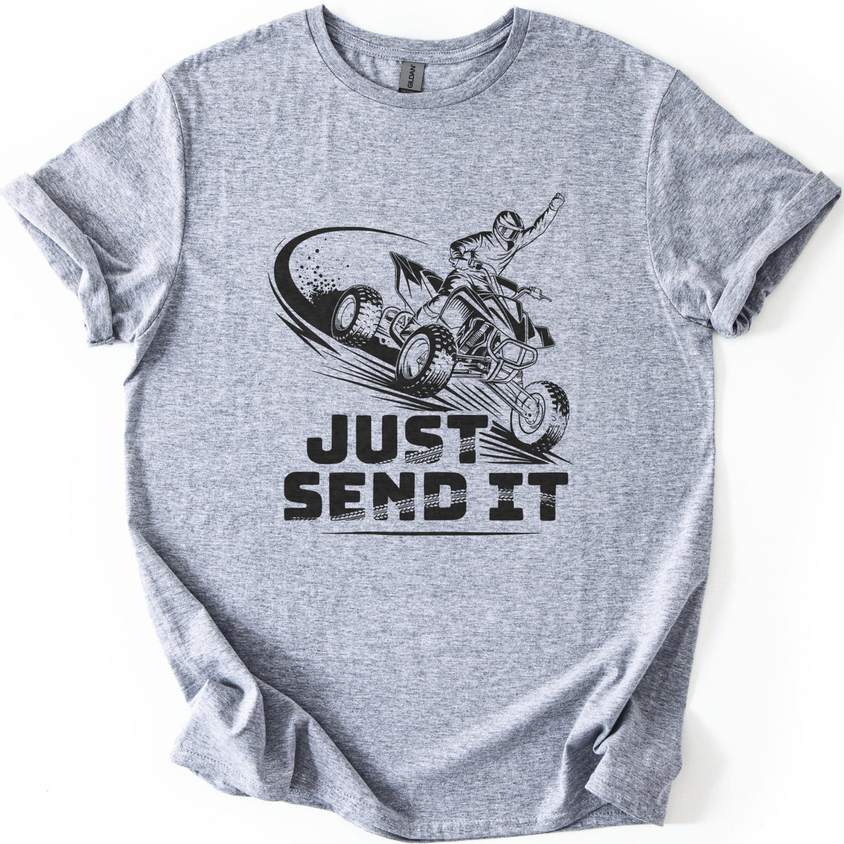 ATV Just Send It Comfort Colors T-shirt in Sport Grey, laid flat. A black drawing of a helmeted rider jumping a quad ATV with one fist in the air and a dust trail behind, above the words Just Send It in tire-tread lettering. Sleeves are cuffed, on a white surface.