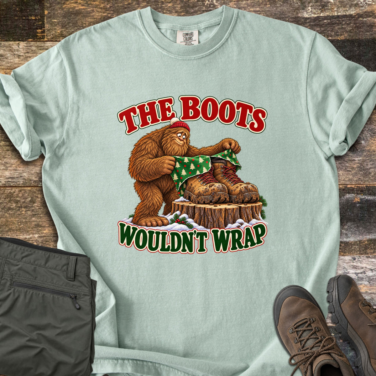 The Boots Wouldn't Wrap Comfort Colors T-shirt in Bay, laid flat. A Bigfoot in a red beanie tries to wrap muddy hiking boots on a snowy stump in green Christmas tree paper, between the words The Boots Wouldn't Wrap. Olive pants and brown hiking boots lie beside it on weathered wood.