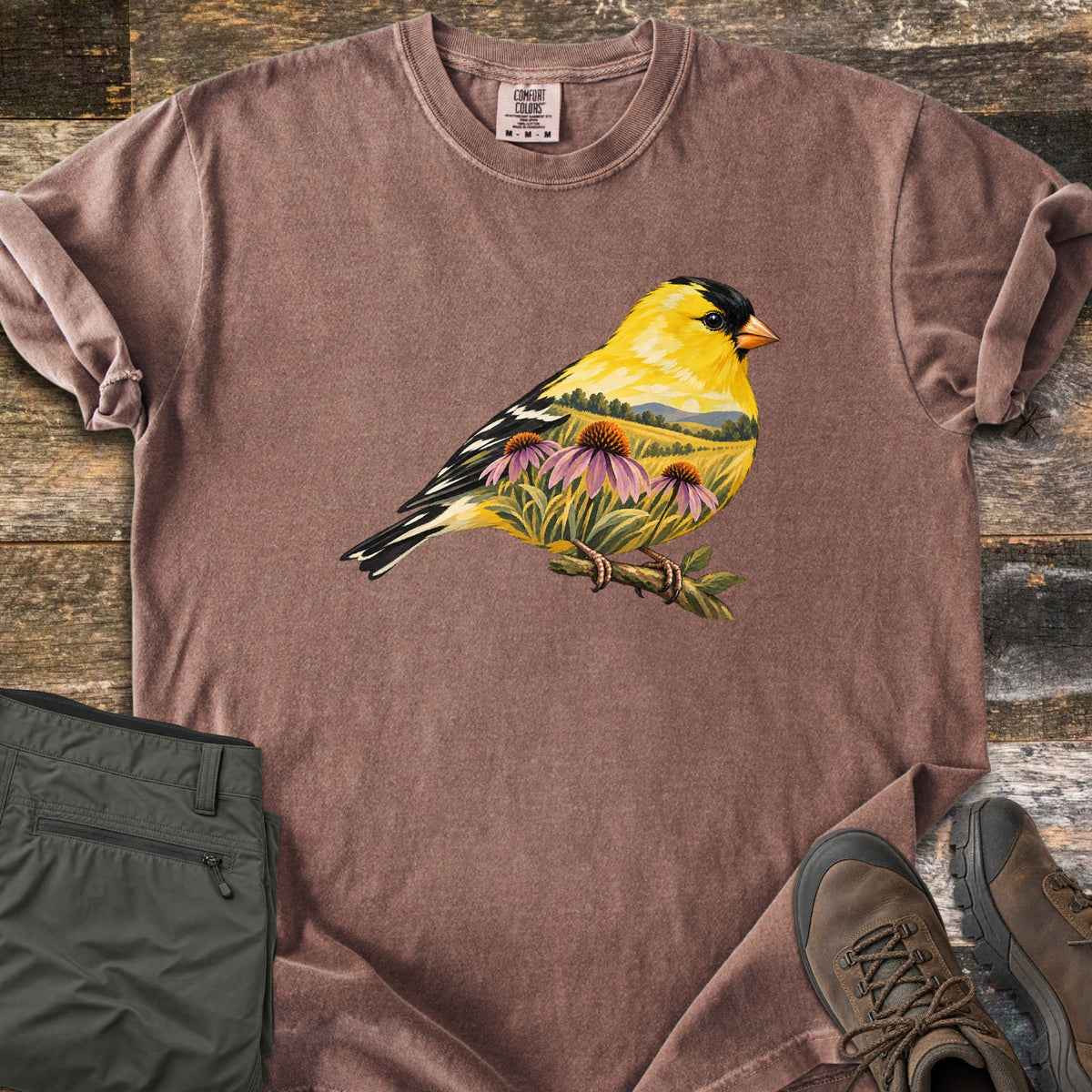 Goldfinch Meadow Comfort Colors T-shirt in Espresso, laid flat. A goldfinch on a twig, its yellow body holding a painted meadow with purple coneflowers, green hills and a setting sun. No text. Folded olive cargo pants and brown hiking boots sit on rustic wood planks.