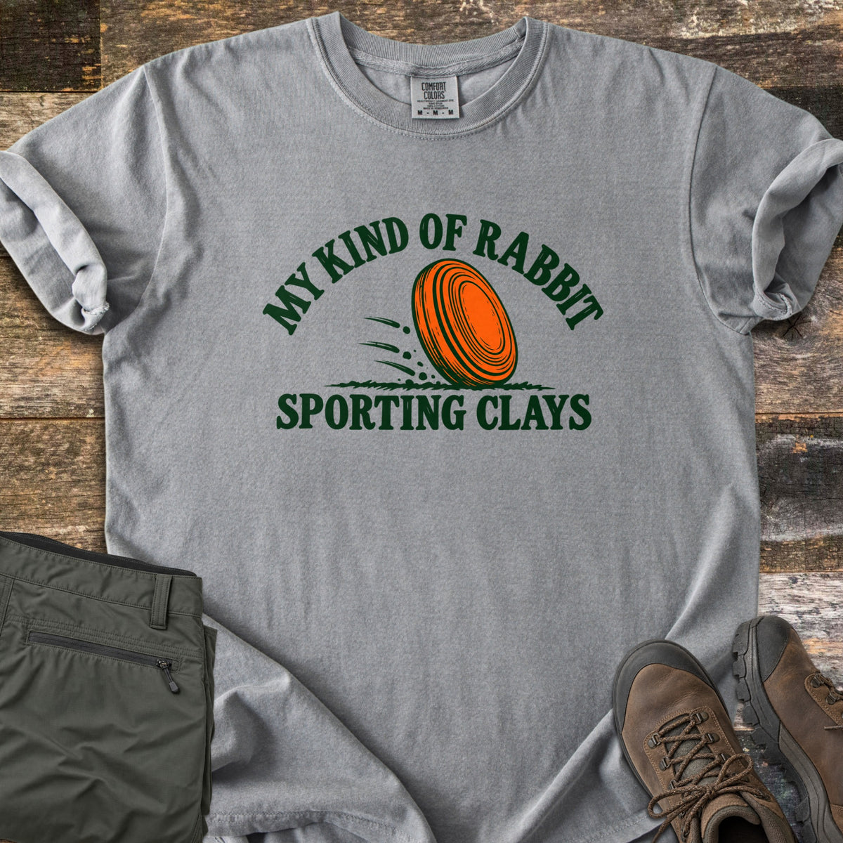 My Kind of Rabbit Comfort Colors T-shirt in Grey, laid flat. An orange clay target rolls through grass between the curved words My Kind of Rabbit and Sporting Clays in dark green. Olive hiking pants and brown boots lie beside it on weathered wood planks.