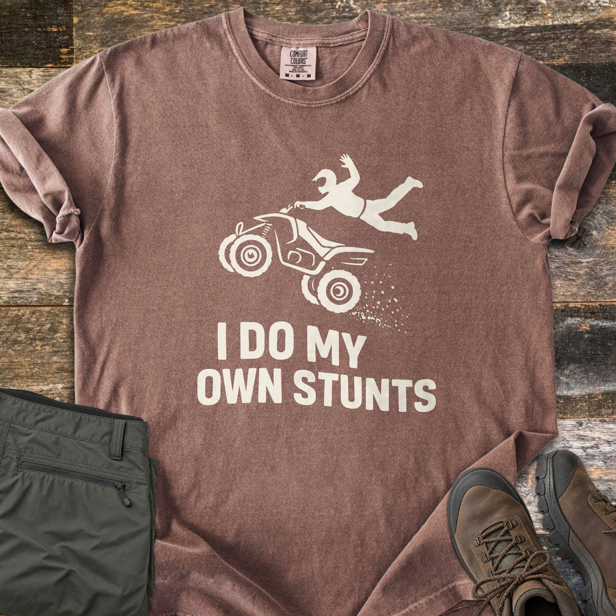 ATV Stunts Comfort Colors T-shirt in Espresso, laid flat. A white silhouette of a helmeted rider flying off a wheelie-popping ATV kicking up dirt, above the words I Do My Own Stunts. Olive cargo pants and brown hiking boots sit beside it on weathered wood.