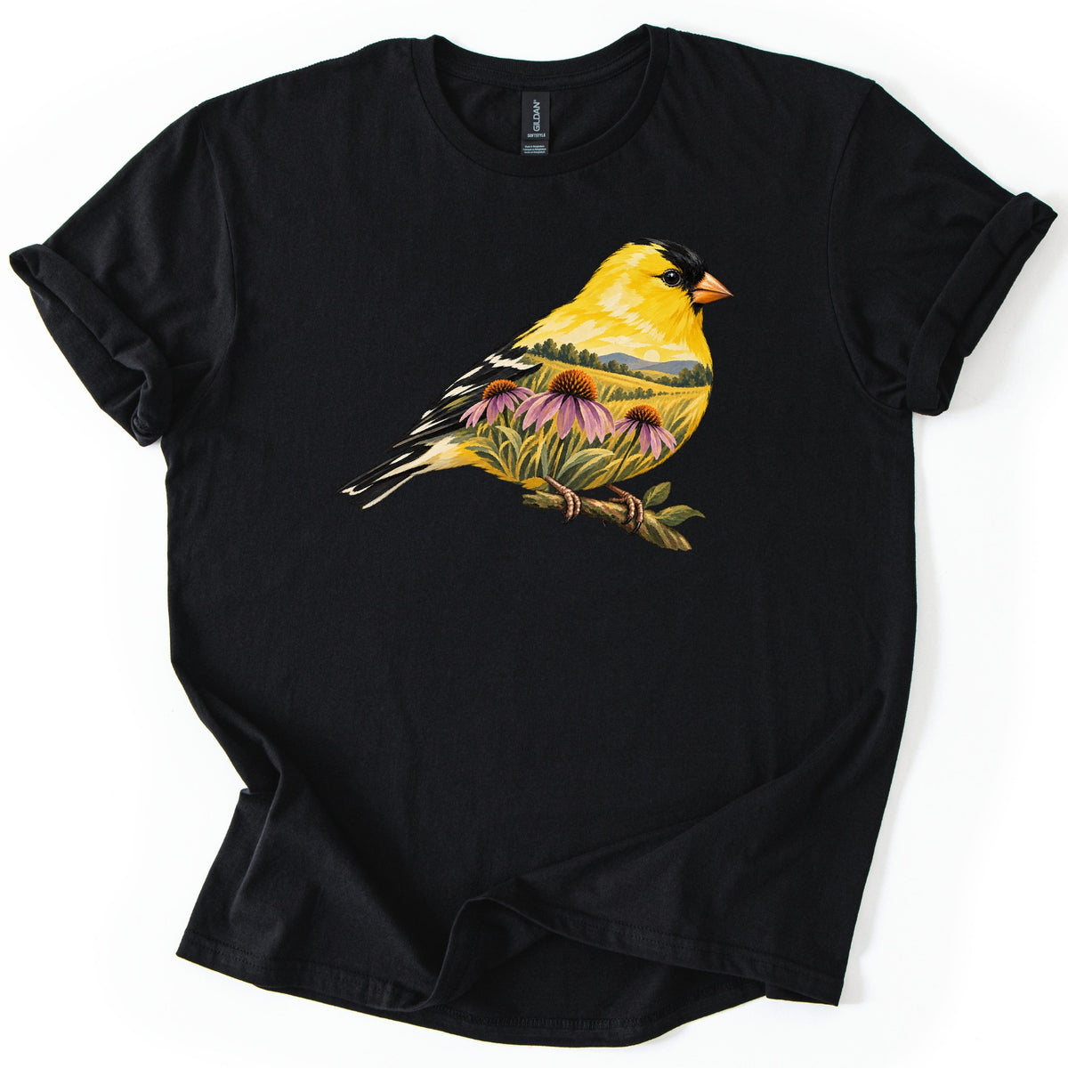 Goldfinch Meadow Comfort Colors T-shirt in Black, laid flat. A yellow goldfinch with black cap and striped wings sits on a twig, its body showing a painted meadow with pink coneflowers, green grass and rolling hills. No text. Laid on a white surface.