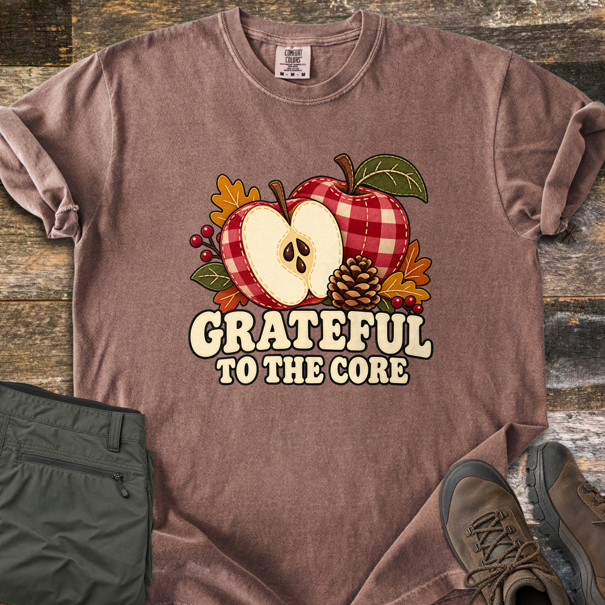 Grateful to the Core Comfort Colors T-shirt in Espresso, laid flat. A red gingham apple and an apple half with seeds sit with a pinecone, autumn leaves and berries above the words Grateful to the Core. Sleeves rolled, it lies on weathered wood by olive pants and brown boots.