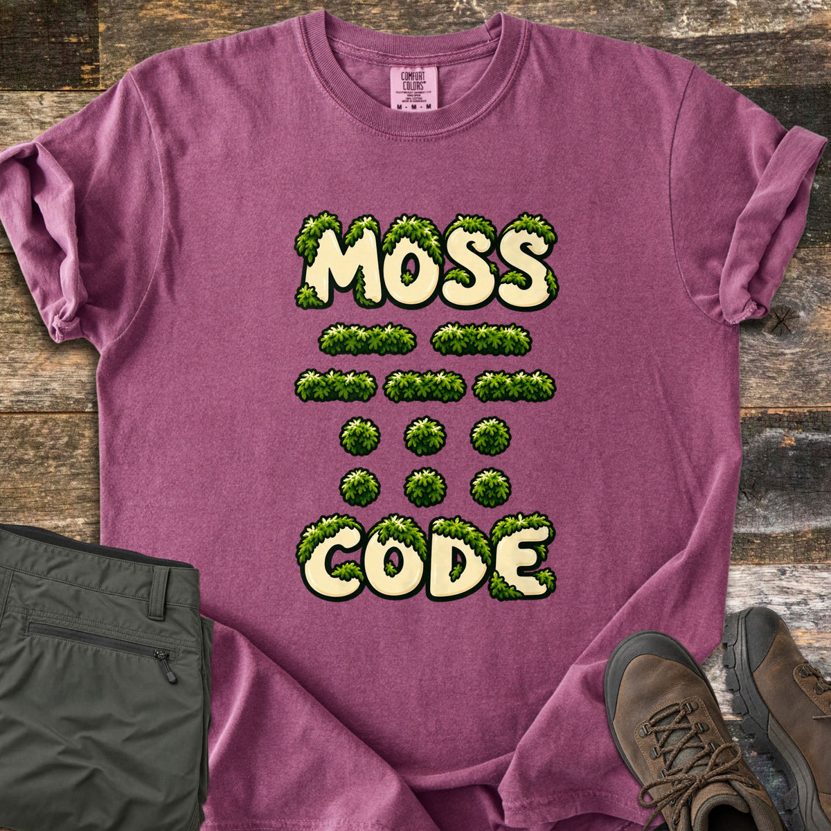 Moss Code Comfort Colors T-shirt in Berry, laid flat. Moss-covered cream bubble letters spell MOSS, then moss-tuft dashes and dots spelling MOSS in Morse code, then CODE. It lies on weathered wood beside folded olive hiking pants and a brown hiking boot.