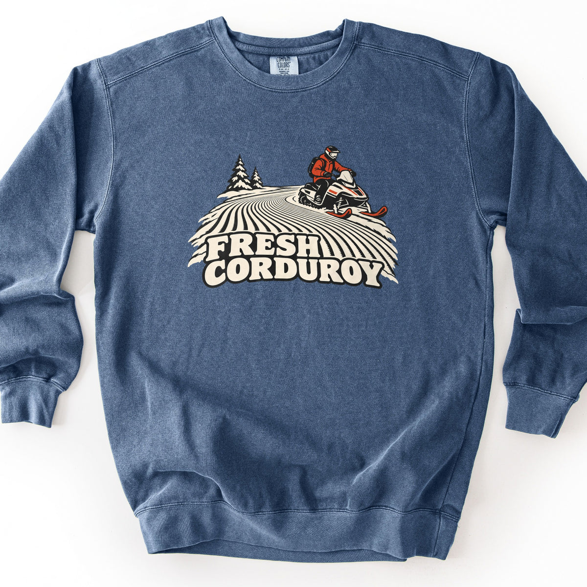Fresh Corduroy Comfort Colors crewneck sweatshirt in Denim, laid flat. A rider in a red jacket and helmet drives a snowmobile over a groomed, striped snow trail with pine trees behind, above the words Fresh Corduroy. The sweatshirt lies on a white background.