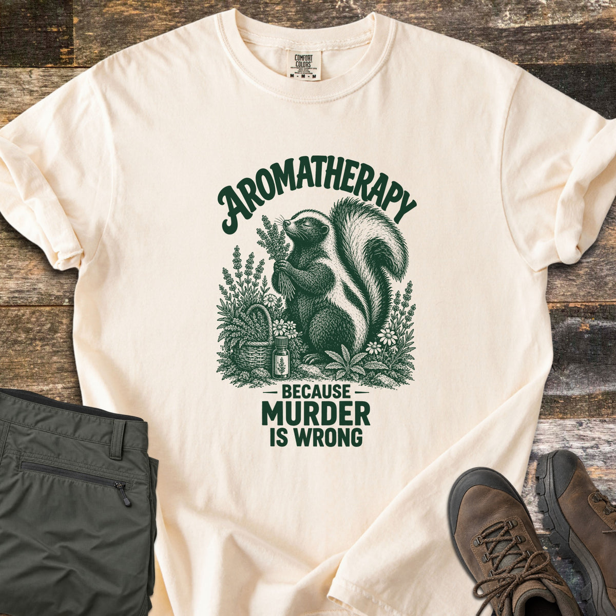 Aromatherapy Because Murder Is Wrong Comfort Colors T-shirt in Ivory, laid flat. An engraving-style skunk sniffs a bundle of lavender among flowers, a basket and an oil bottle, in dark green ink, with the words Aromatherapy, Because Murder Is Wrong. Olive pants and brown hiking boots lie on wood planks.
