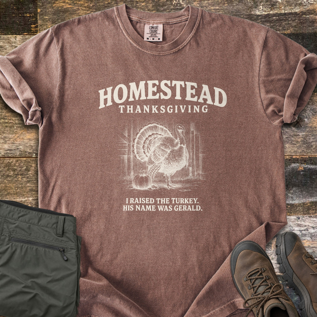Homestead Thanksgiving Comfort Colors T-shirt in Espresso, laid flat. Cream print of a fanned-tail turkey by a barn door and pumpkin, under the words Homestead Thanksgiving, above I Raised the Turkey. His Name Was Gerald. On weathered wood with olive pants and brown hiking boots.