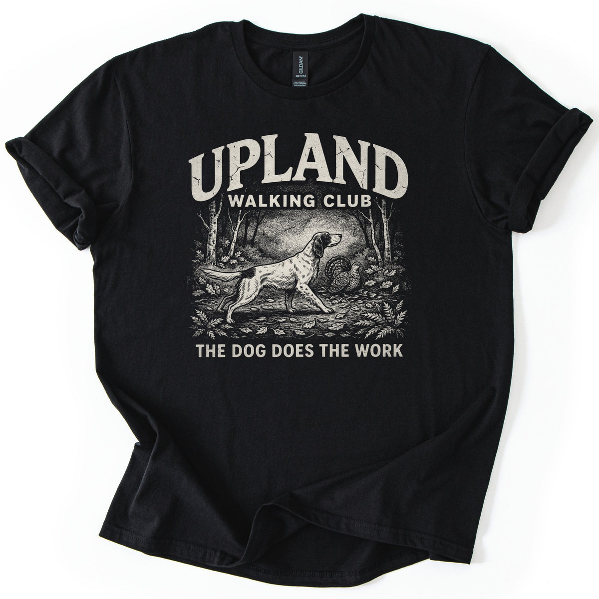Upland Walking Club Comfort Colors T-shirt in Black, laid flat. A cream engraved-style print shows a setter dog pointing toward a grouse among trees and leaves, under Upland Walking Club and over The Dog Does The Work. The shirt lies on white with rolled sleeves.