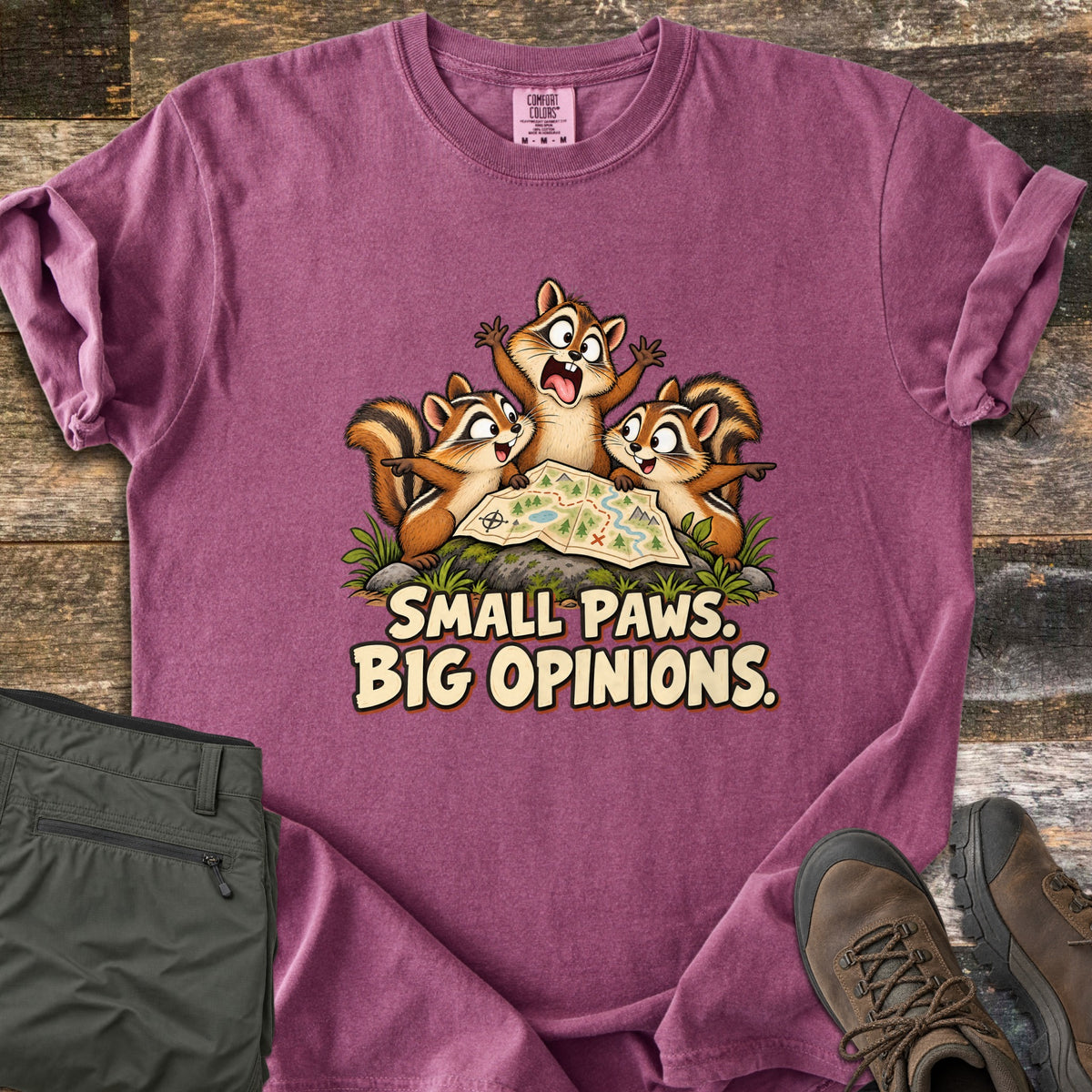 Small Paws. Big Opinions. Comfort Colors T-shirt in Berry, laid flat. Three cartoon chipmunks squabble over a trail map on a mossy rock, the middle one shouting, above the words SMALL PAWS. BIG OPINIONS. Olive hiking pants and brown hiking boots lie beside it on rustic wood.