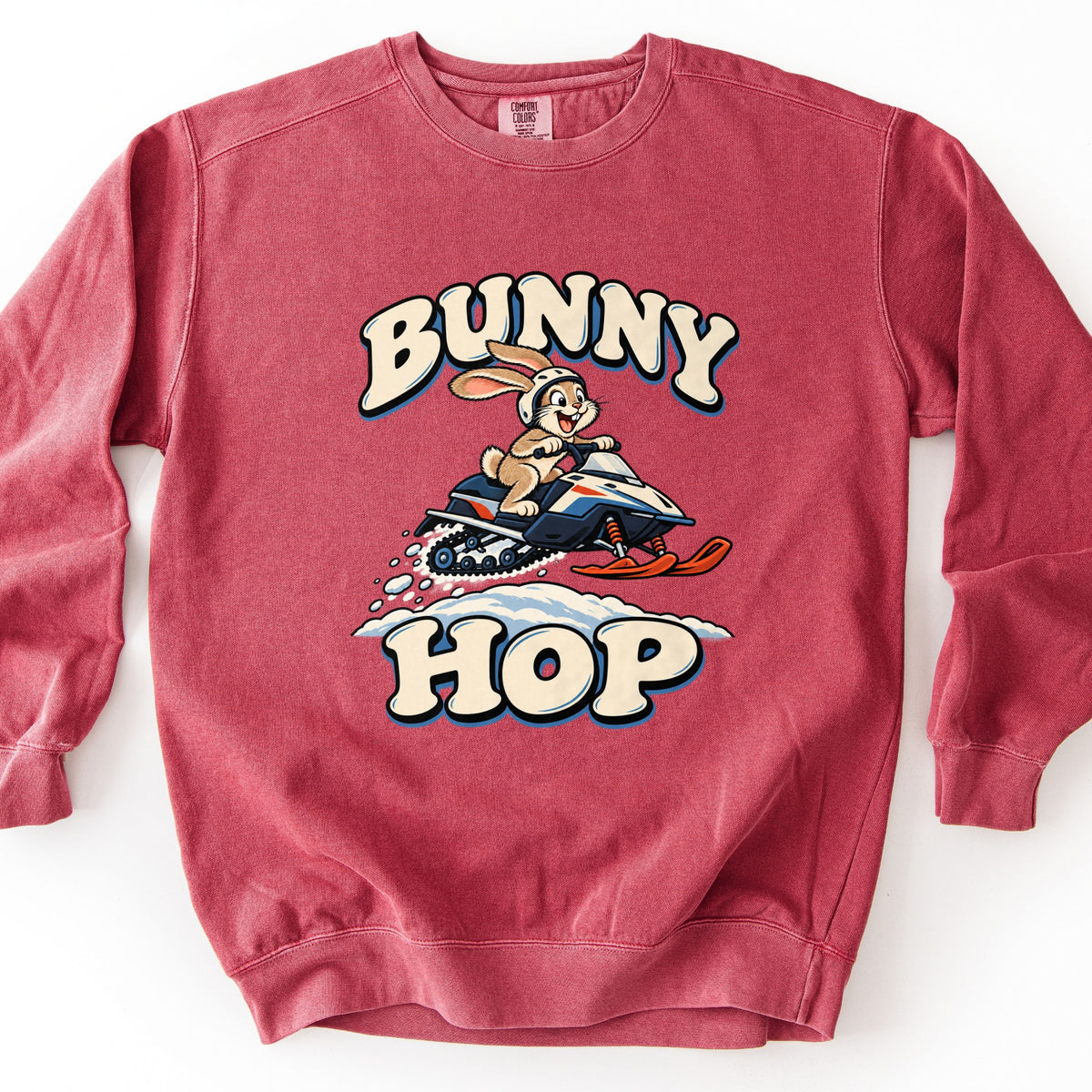 Bunny Hop Comfort Colors crewneck sweatshirt in Crimson, laid flat. A grinning cartoon bunny in a white helmet rides a navy, white and orange snowmobile over a snowbank between the words Bunny Hop in cream bubble letters. The sweatshirt lies on a white surface.