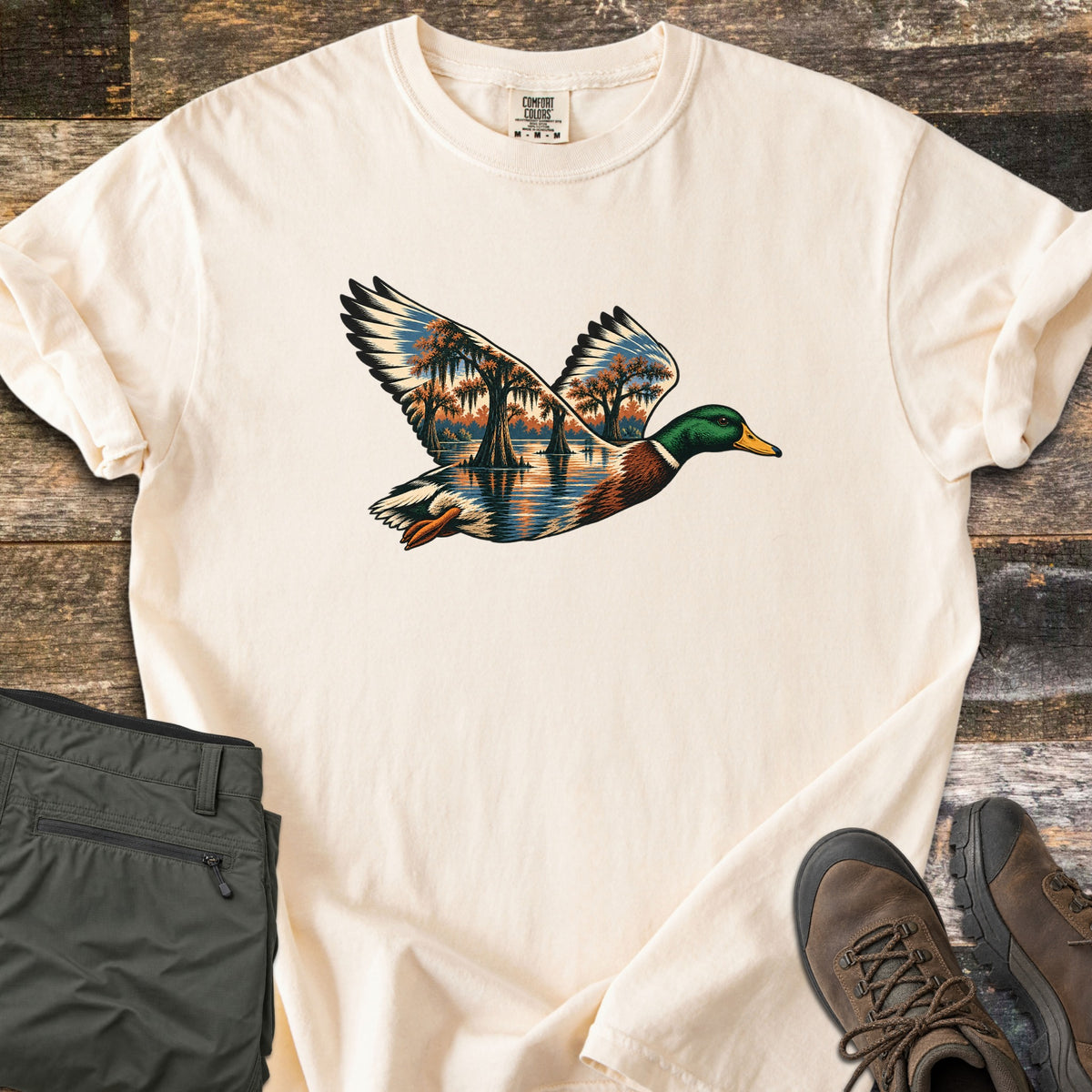 Mallard Flooded Oaks Comfort Colors T-shirt in Ivory, laid flat. A flying mallard drake whose wings and body are filled with a scene of moss-draped oaks standing in still water at sunset. Laid on weathered wood beside folded olive pants and brown hiking boots.