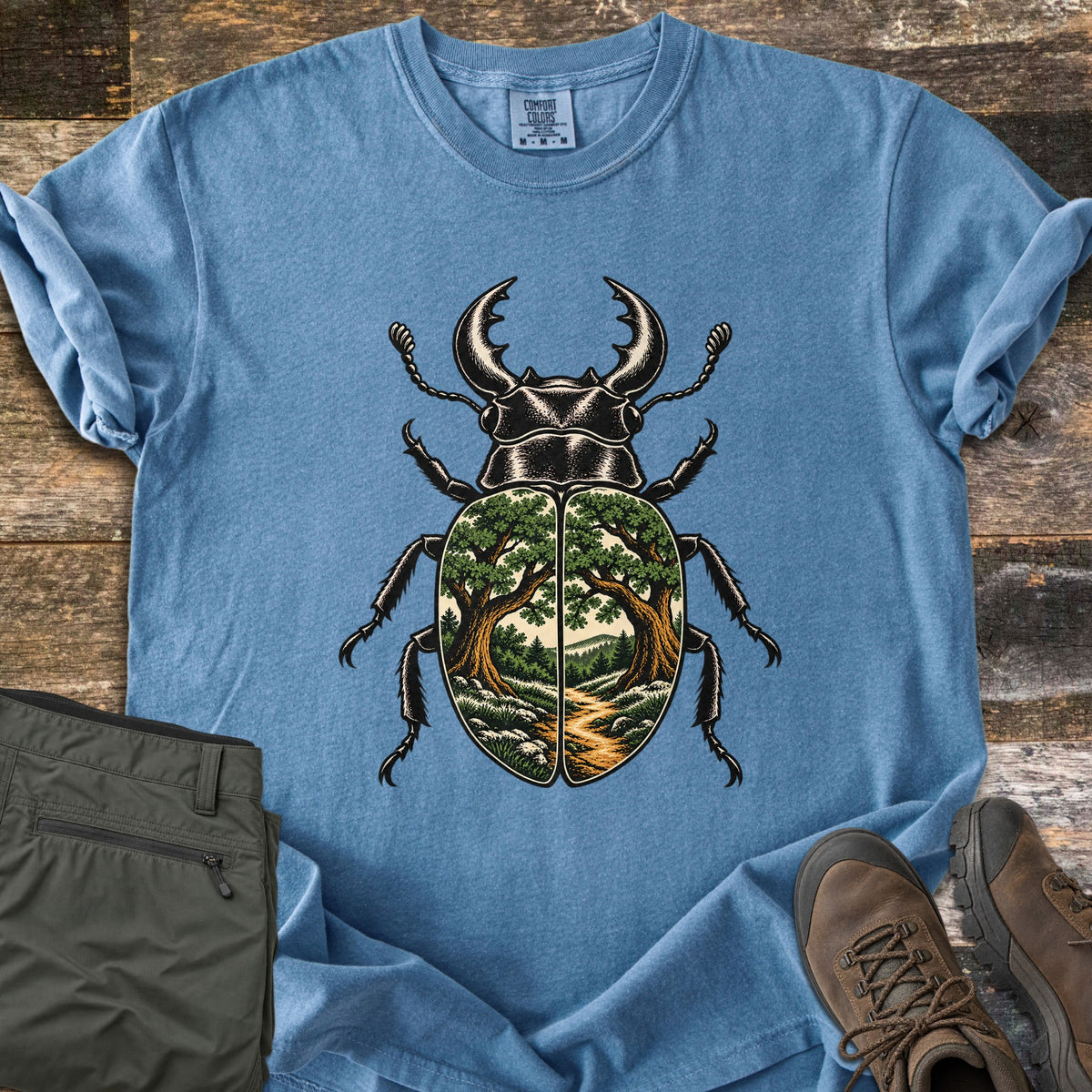 Beetle Forest Path Comfort Colors T-shirt in Blue Jean, laid flat. A black stag beetle whose wing covers show a forest scene of two oak trees framing a dirt path toward distant hills. Folded olive hiking pants and brown hiking boots sit beside it on weathered wood planks.