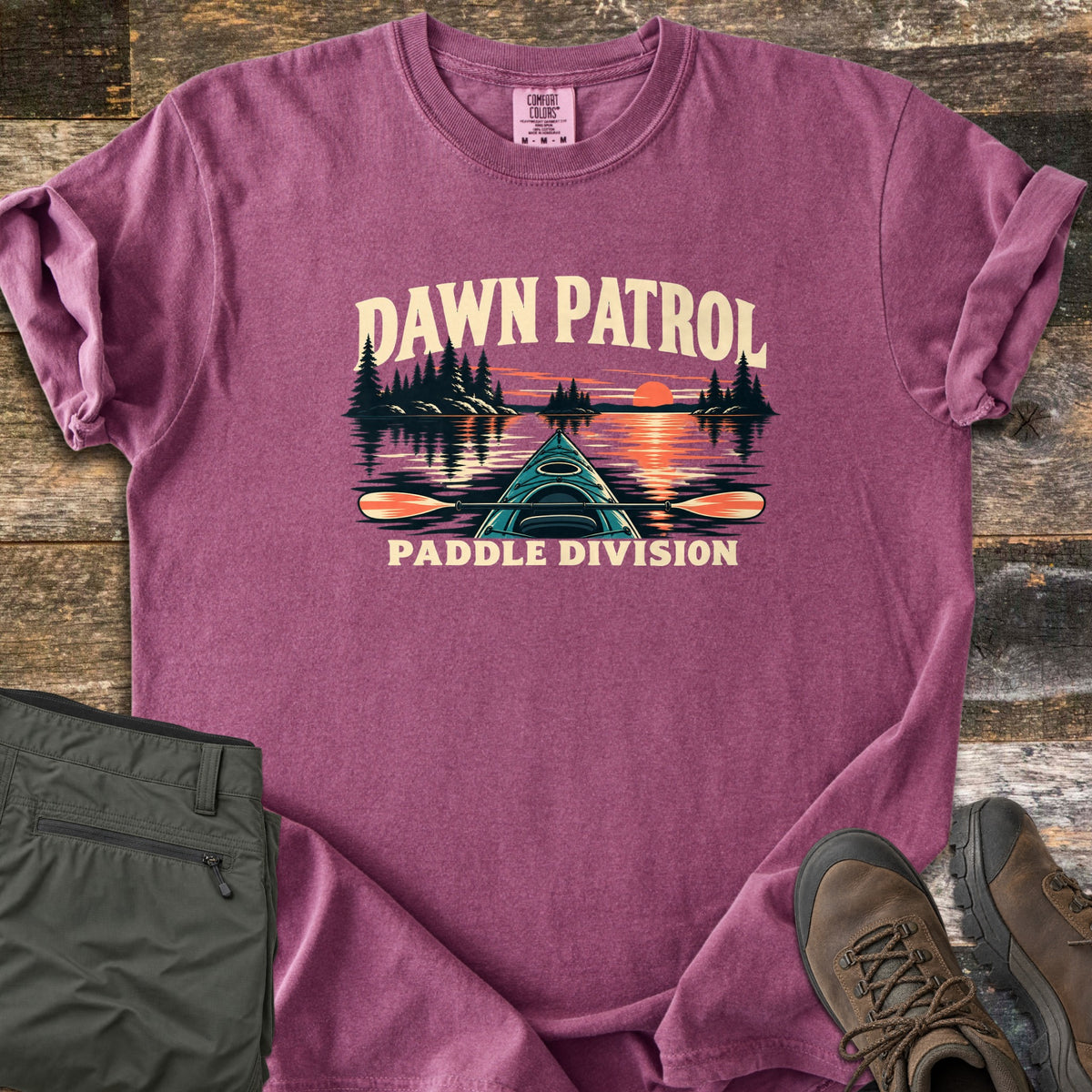 Dawn Patrol Paddle Division Comfort Colors T-shirt in Berry, laid flat. The words Dawn Patrol arch over a teal kayak bow and paddle on a lake with pine islands and an orange sunrise, above the words Paddle Division. Olive hiking pants and brown hiking boots lie beside it on weathered wood.