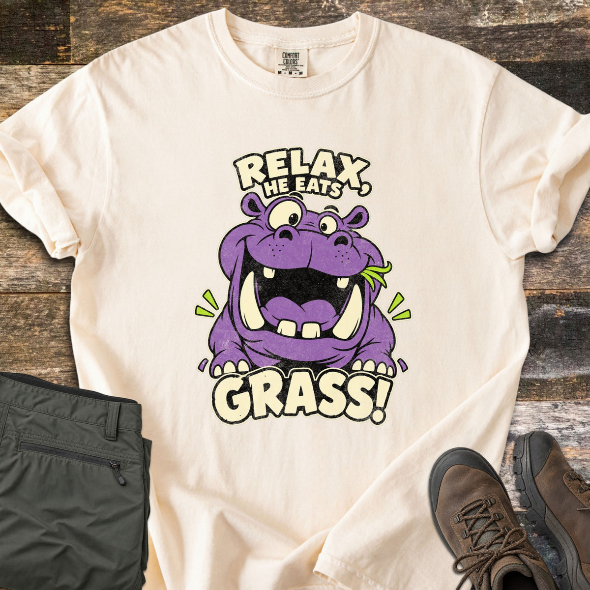 Relax He Eats Grass Comfort Colors T-shirt in Ivory, laid flat. A grinning purple cartoon hippo with a sprig of green grass in its mouth sits between the words Relax, He Eats and Grass! Folded olive pants and brown hiking boots lie beside it on weathered wood planks.