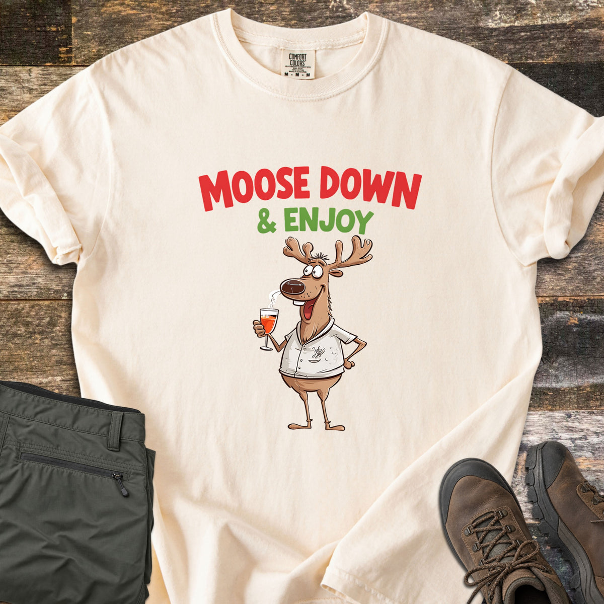 Moose Down And Enjoy Comfort Colors T-shirt in Ivory, laid flat. A cartoon moose in a white shirt holds a glass of orange drink under the words Moose Down & Enjoy in red and green. Shown on rustic wood with folded olive pants and brown hiking boots.