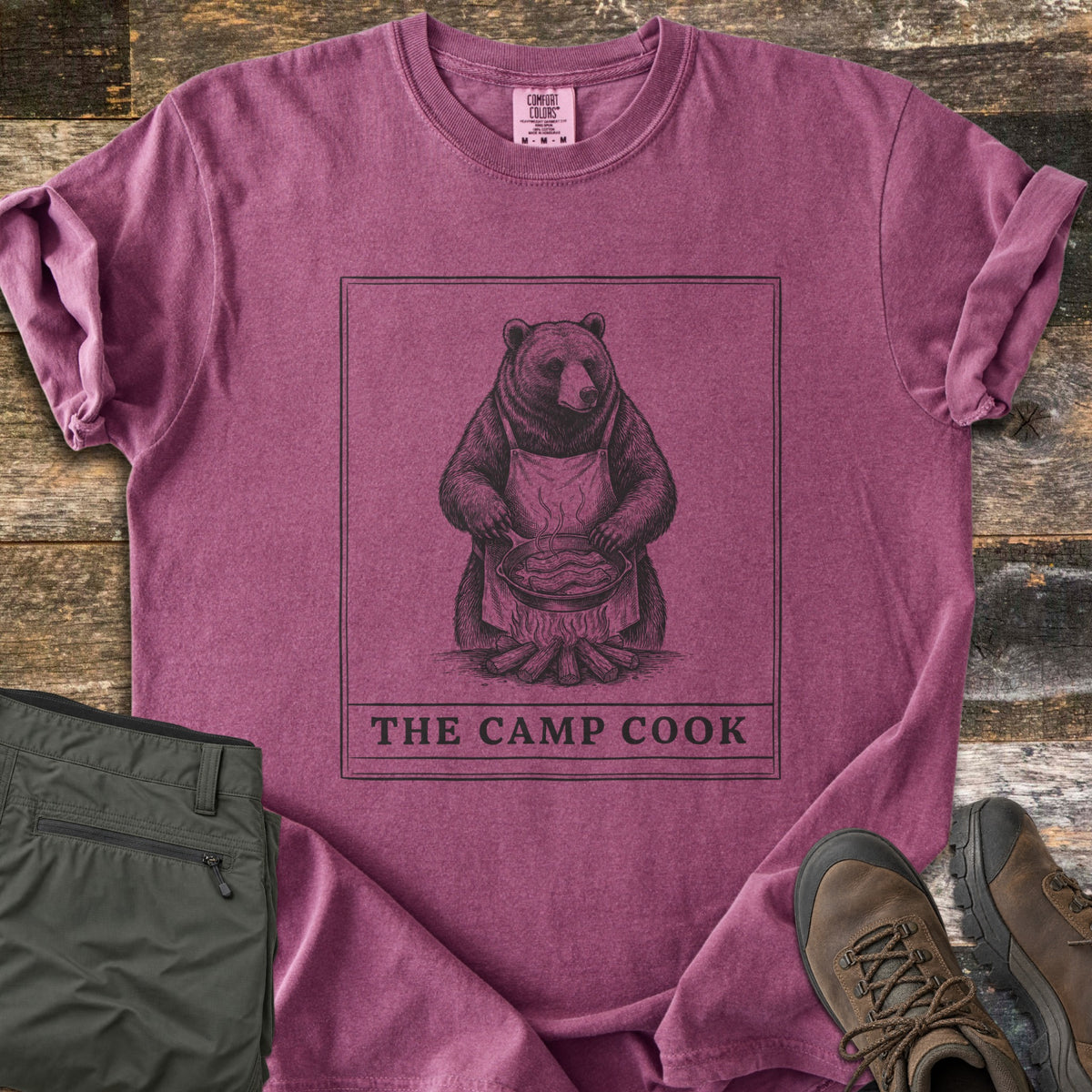 The Camp Cook Comfort Colors T-shirt in Berry, laid flat. A framed sketch of a bear in an apron frying bacon in a skillet over a campfire, above the words THE CAMP COOK. Sleeves are cuffed on weathered wood beside olive hiking pants and brown hiking boots.