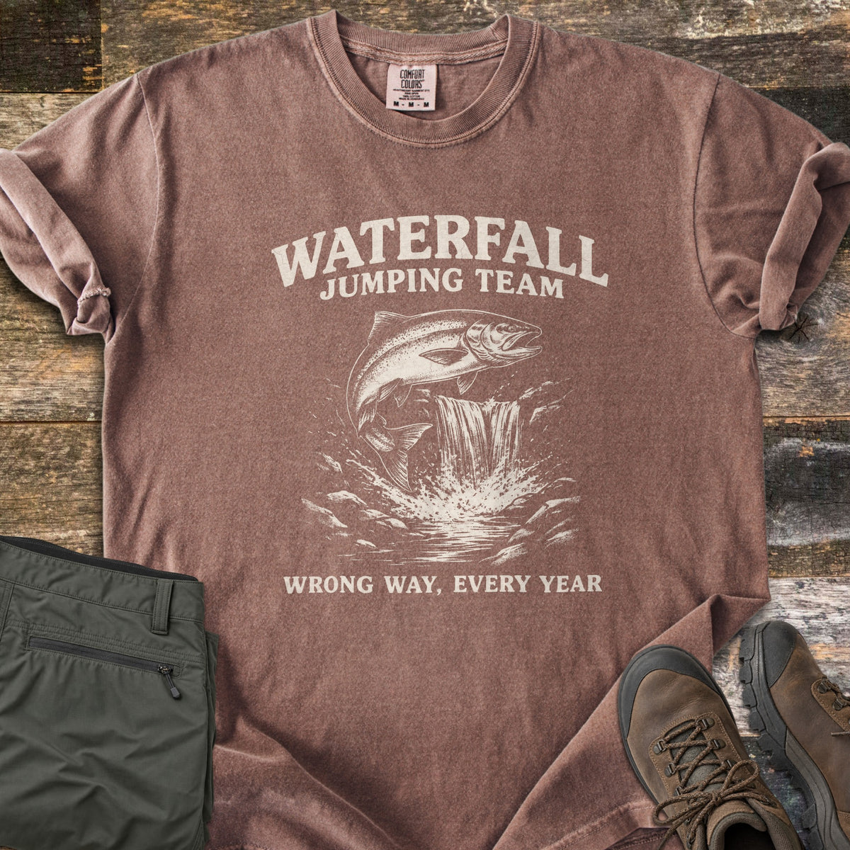Waterfall Jumping Team Comfort Colors T-shirt in Espresso, laid flat. A salmon leaps beside a small waterfall between the words Waterfall Jumping Team and Wrong Way, Every Year, printed in cream. Shown on rustic wood with folded olive pants and brown hiking boots.
