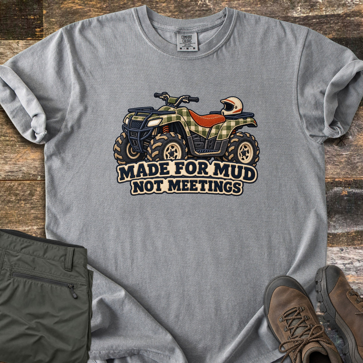 Made for Mud Not Meetings Comfort Colors T-shirt in Grey, laid flat. A green plaid ATV with an orange seat, muddy tires and a white helmet on the rack sits above the words Made for Mud Not Meetings. It lies on weathered wood beside folded olive pants and brown hiking boots.
