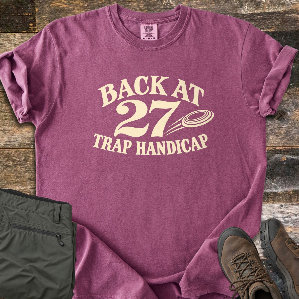 Back at 27 Trap Handicap Comfort Colors T-shirt in Berry, laid flat. Cream print reads Back at 27 in an arch above Trap Handicap, with a clay target flying beside the 27. Sleeves rolled on a rustic wood surface, with olive cargo pants and brown hiking boots.
