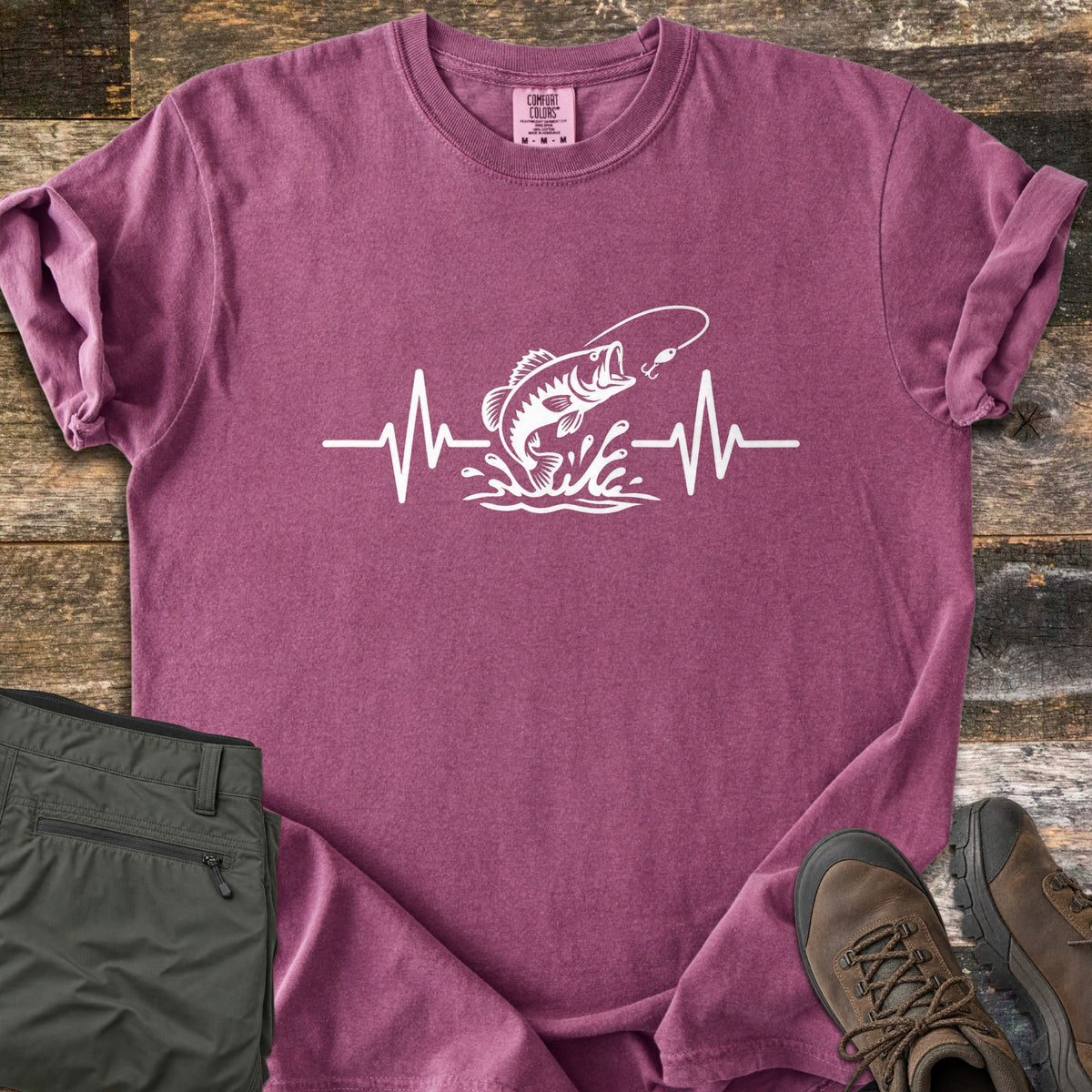 Fishing Heartbeat Comfort Colors T-shirt in Berry, laid flat. A white print of a largemouth bass leaping from splashing water toward a fishing lure, set in the middle of a heartbeat line. Sleeves are rolled, on rustic wood with olive cargo shorts and brown hiking boots.
