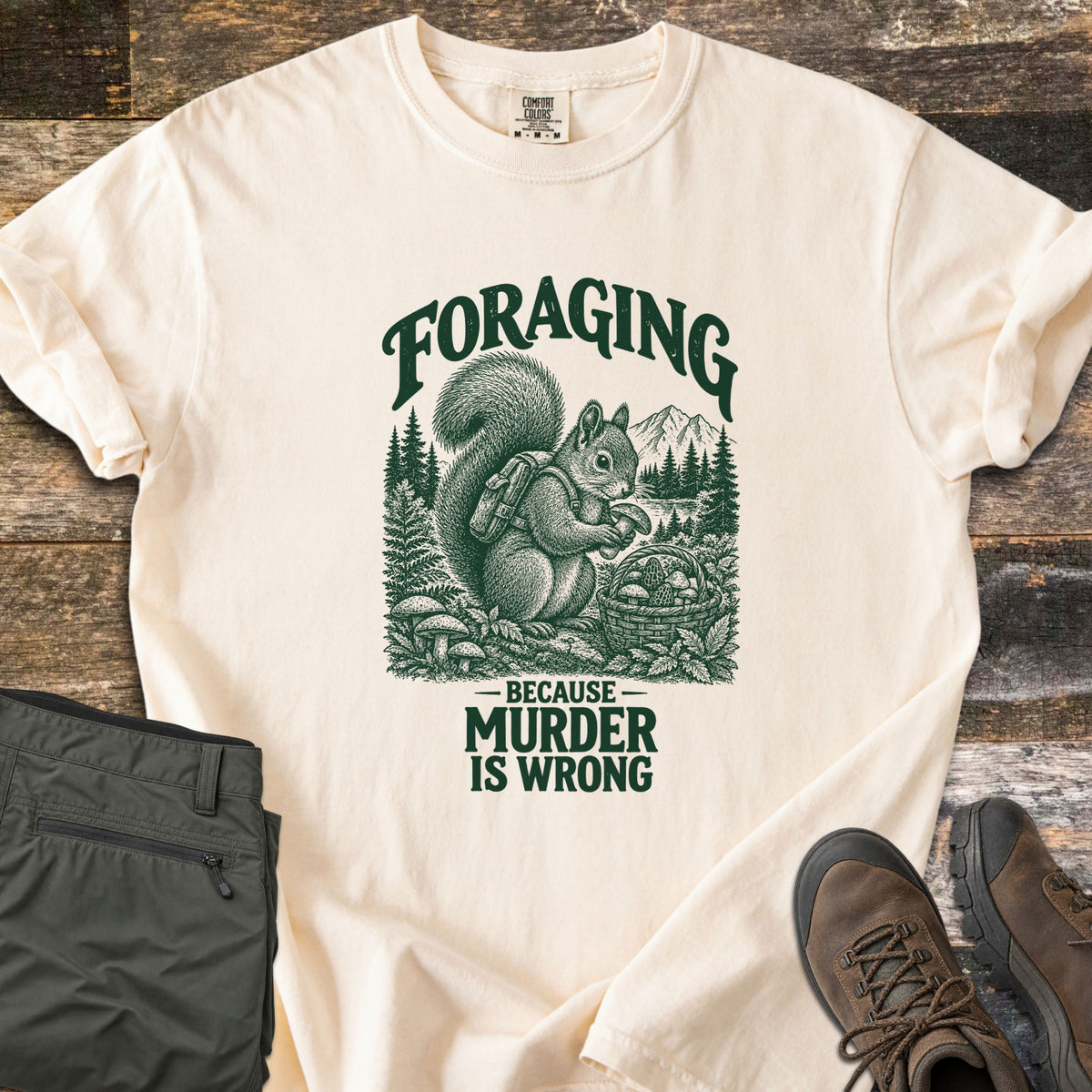 Foraging Murder Squirrel Comfort Colors T-shirt in Ivory, laid flat. Green engraving-style art of a squirrel with a backpack holding a mushroom by a basket of mushrooms, forest and mountains behind, reads Foraging, Because Murder Is Wrong. Green pants and brown boots on wood.