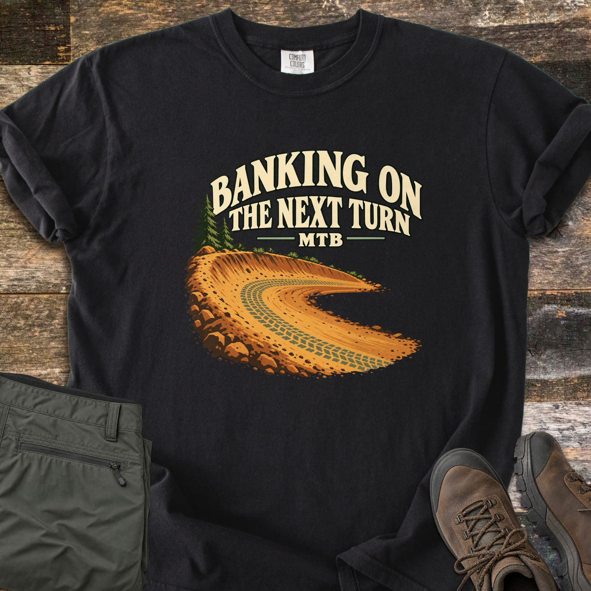 Banking on the Next Turn Comfort Colors T-shirt in Black, laid flat. A dirt berm with tire tracks curves beneath pine trees, under the words Banking on the Next Turn MTB. Sleeves are rolled; olive hiking pants and brown hiking boots sit beside it on weathered wood.