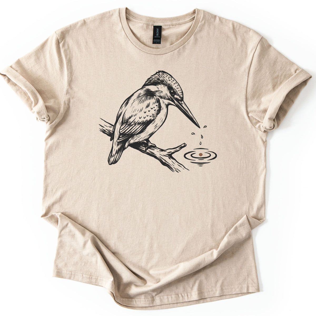 Kingfisher Comfort Colors T-shirt in Sand, laid flat. A black ink drawing of a kingfisher perched on a branch, with droplets falling from its beak into rippling water below. The sleeves are cuffed and the shirt lies on a white background.