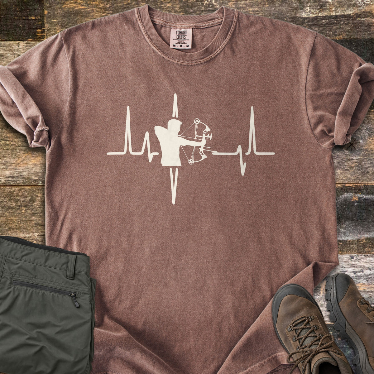 Compound Bow Heartbeat Comfort Colors T-shirt in Espresso, laid flat. A cream heartbeat line with the silhouette of an archer drawing a compound bow at its center. Sleeves are rolled on a wood plank floor, with olive cargo pants and brown hiking boots beside it.