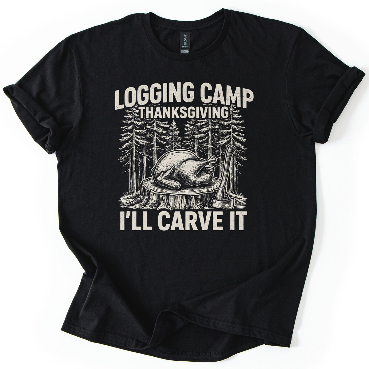 Logging Camp Thanksgiving Comfort Colors T-shirt in Black, laid flat. Cream-colored print of a roast turkey resting on a tree stump with an axe beside it, surrounded by tall pines, under Logging Camp Thanksgiving and above I'll Carve It. The shirt lies on a white surface with cuffed sleeves.