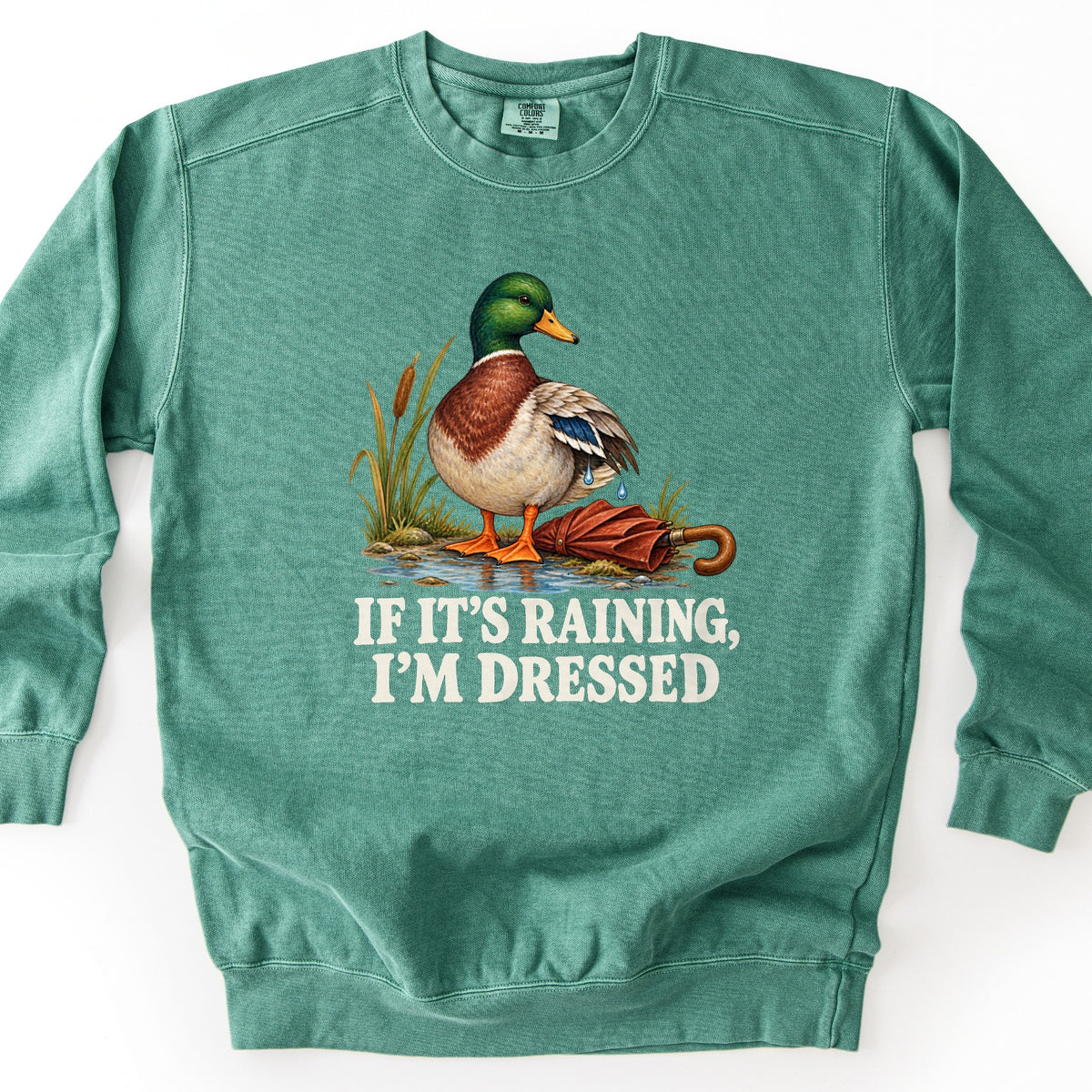 If It's Raining, I'm Dressed Comfort Colors crewneck sweatshirt in Light Green, laid flat. A mallard duck with water dripping from its wing stands in a puddle by cattails beside a closed red umbrella, above the words If It's Raining, I'm Dressed in cream. Shown on a white background.