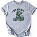 Keep Building Beaver Comfort Colors T-shirt in Sport Grey, laid flat. Dark green sketch-style artwork of a beaver hugging a log while sitting on a pile of cut logs, with Keep Building arched above and No Excuses below. Sleeves are rolled on a white background.